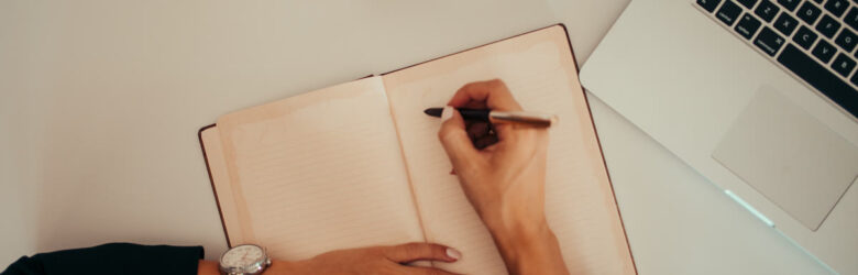 A person taking notes while working on a laptop in a modern workspace — representing a meeting note-taking setup