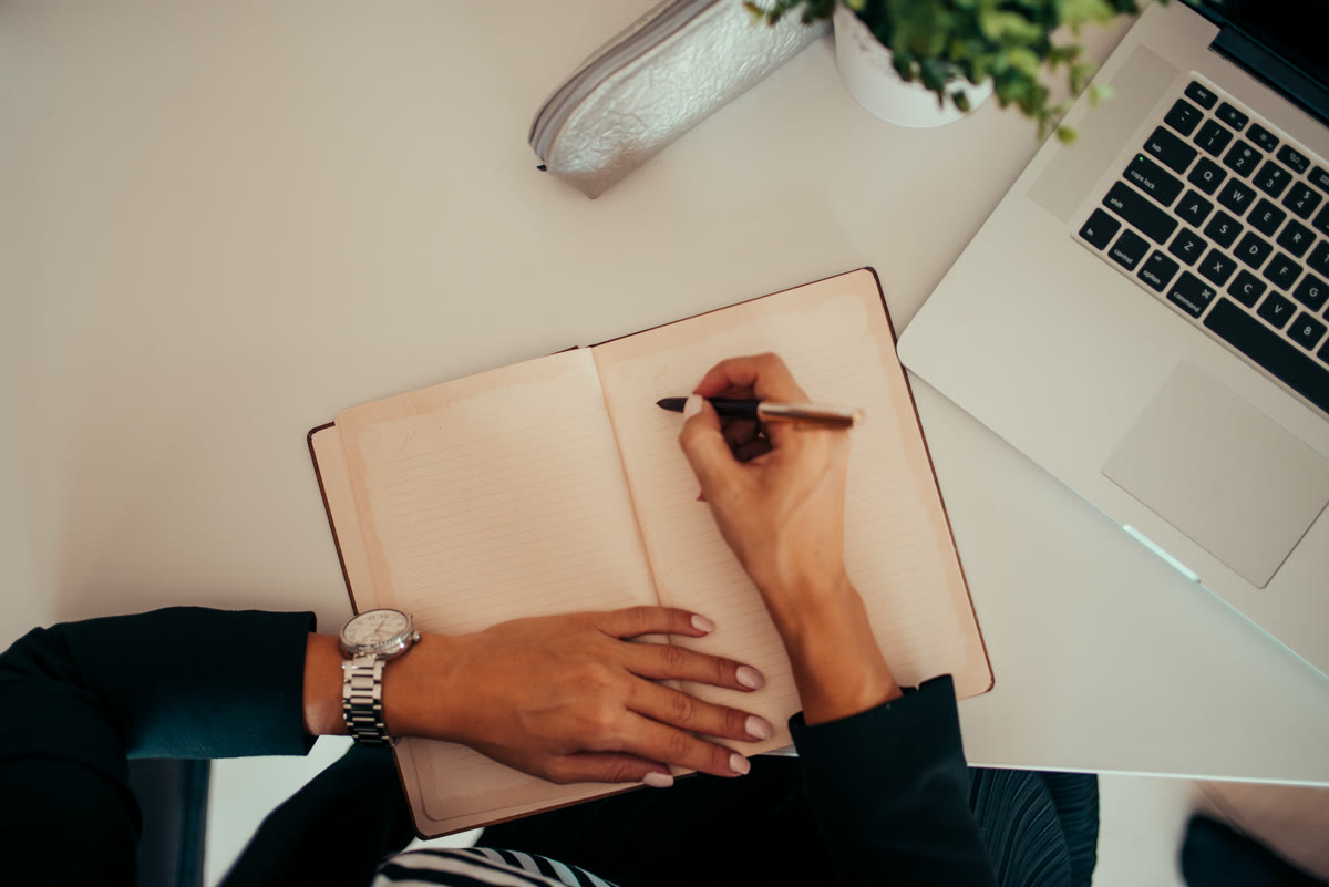 A person taking notes while working on a laptop in a modern workspace