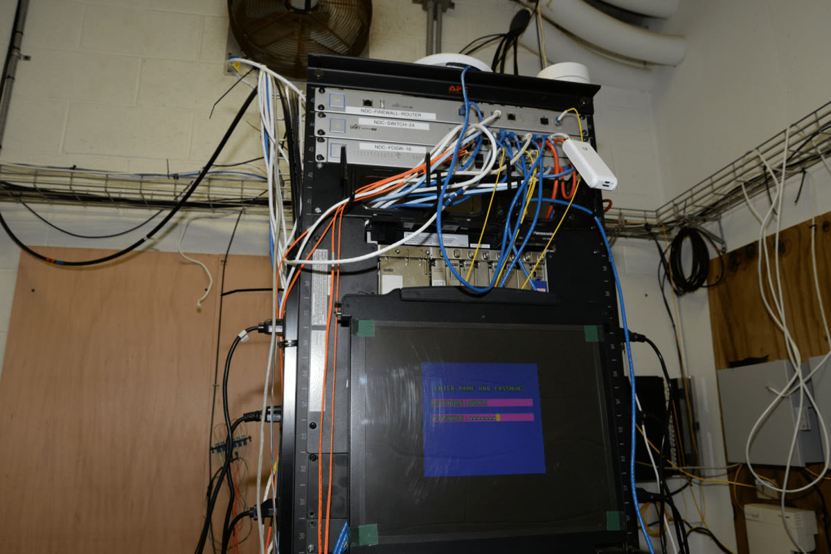 Network server rack with numerous cables and a monitor displaying login credentials on a blue screen